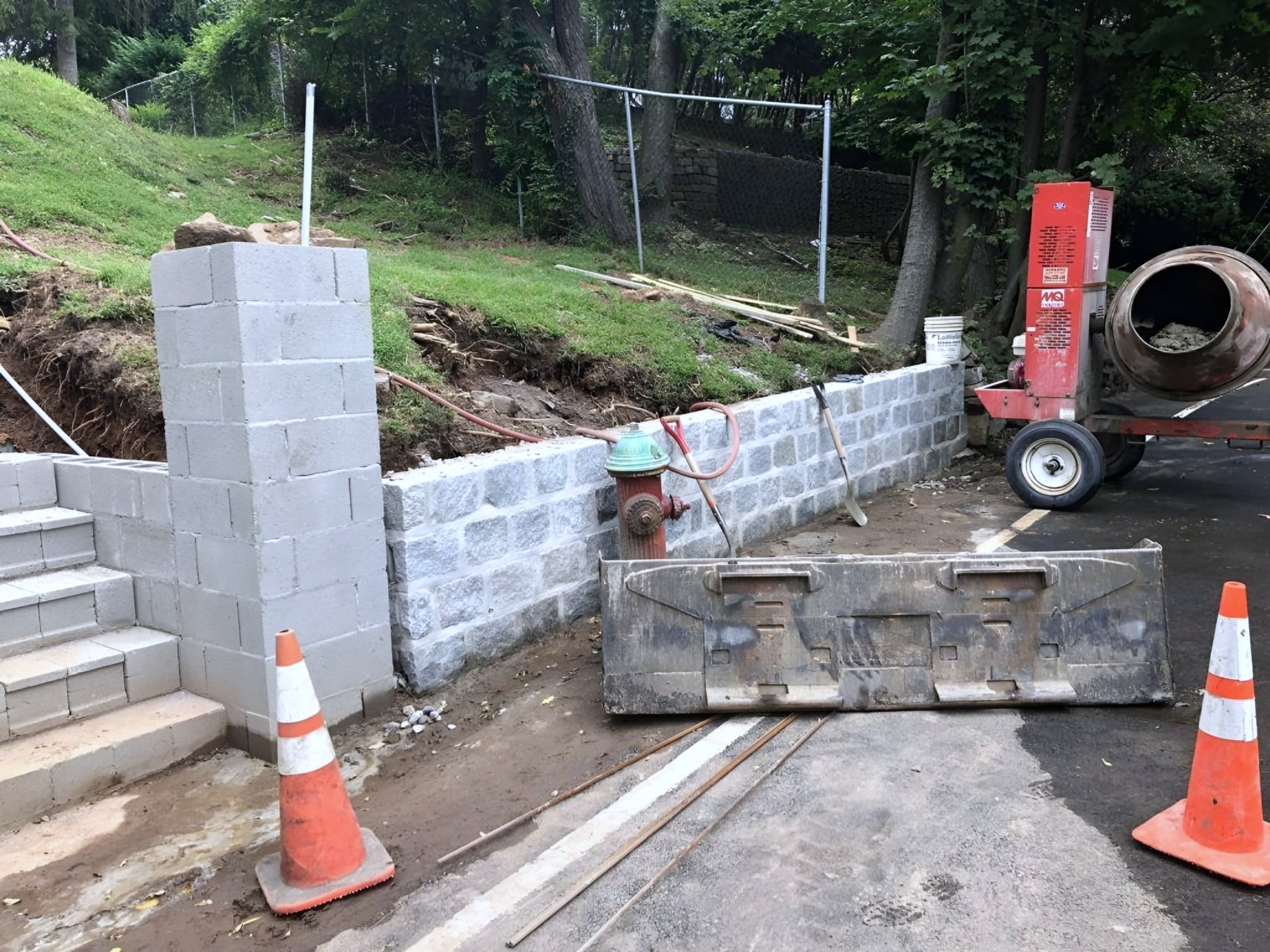 Block retaining wall under construction beside hydrant, cones and cement mixer